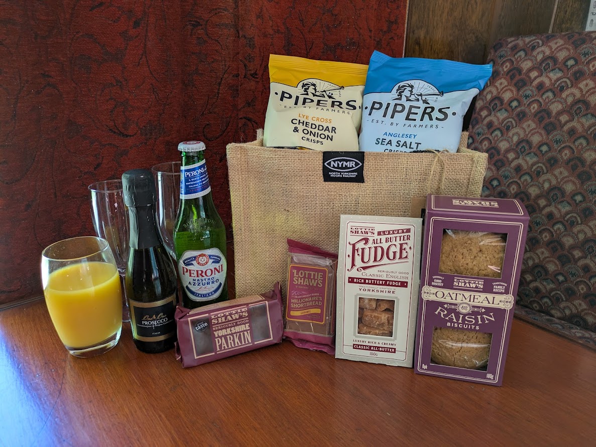 The contents of a travel treats bag including biscuits, cakes, fudge, crisps, Peroni, Prosecco and orange juice.