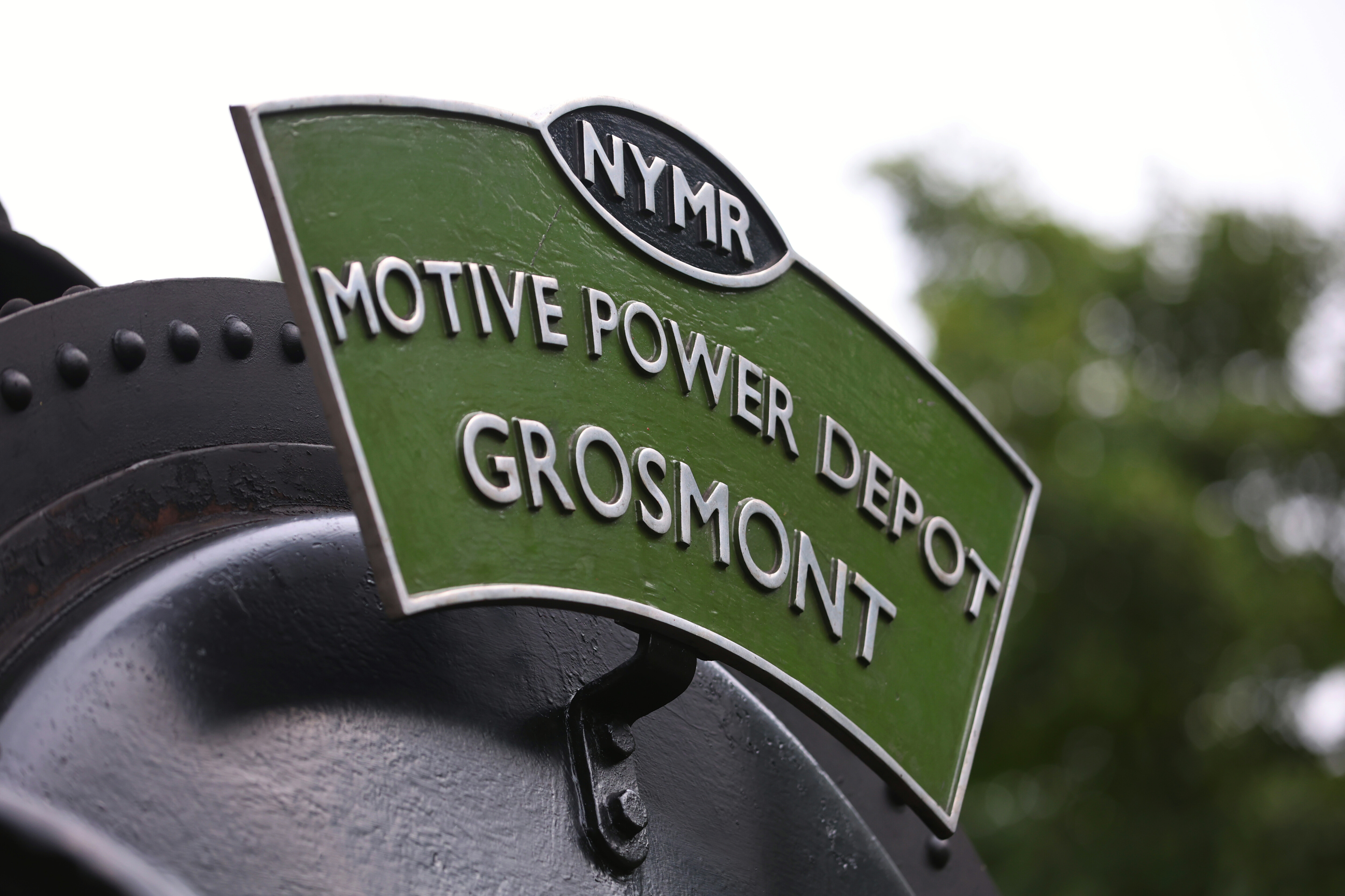 A metal headboard, painted in green and black with the NYMR logo and silver metal text reading Motive Power Deport Grosmont.