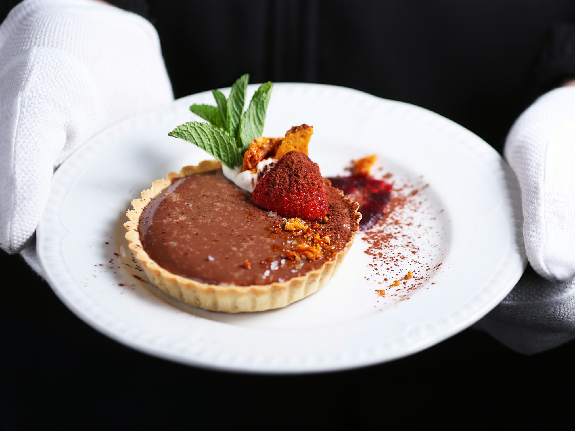 A chocolate tart with fresh strawberry on a plate and held by the white gloved hands of a waiter.