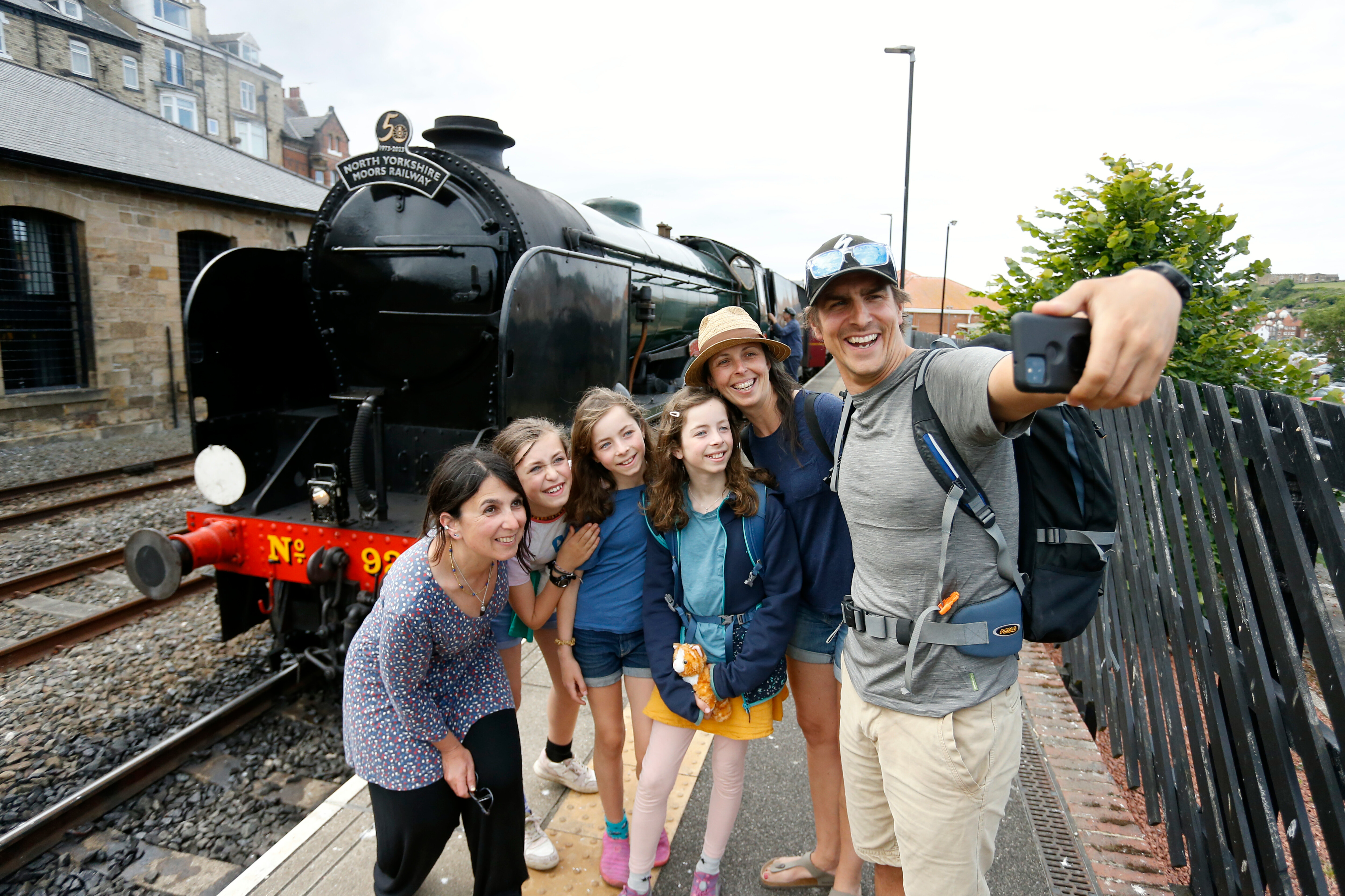A family with three adults and three girls take a selfie at Whitby Station in front of a steam locomotive.