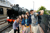 A family with three adults and three girls take a selfie at Whitby Station in front of a steam locomotive.