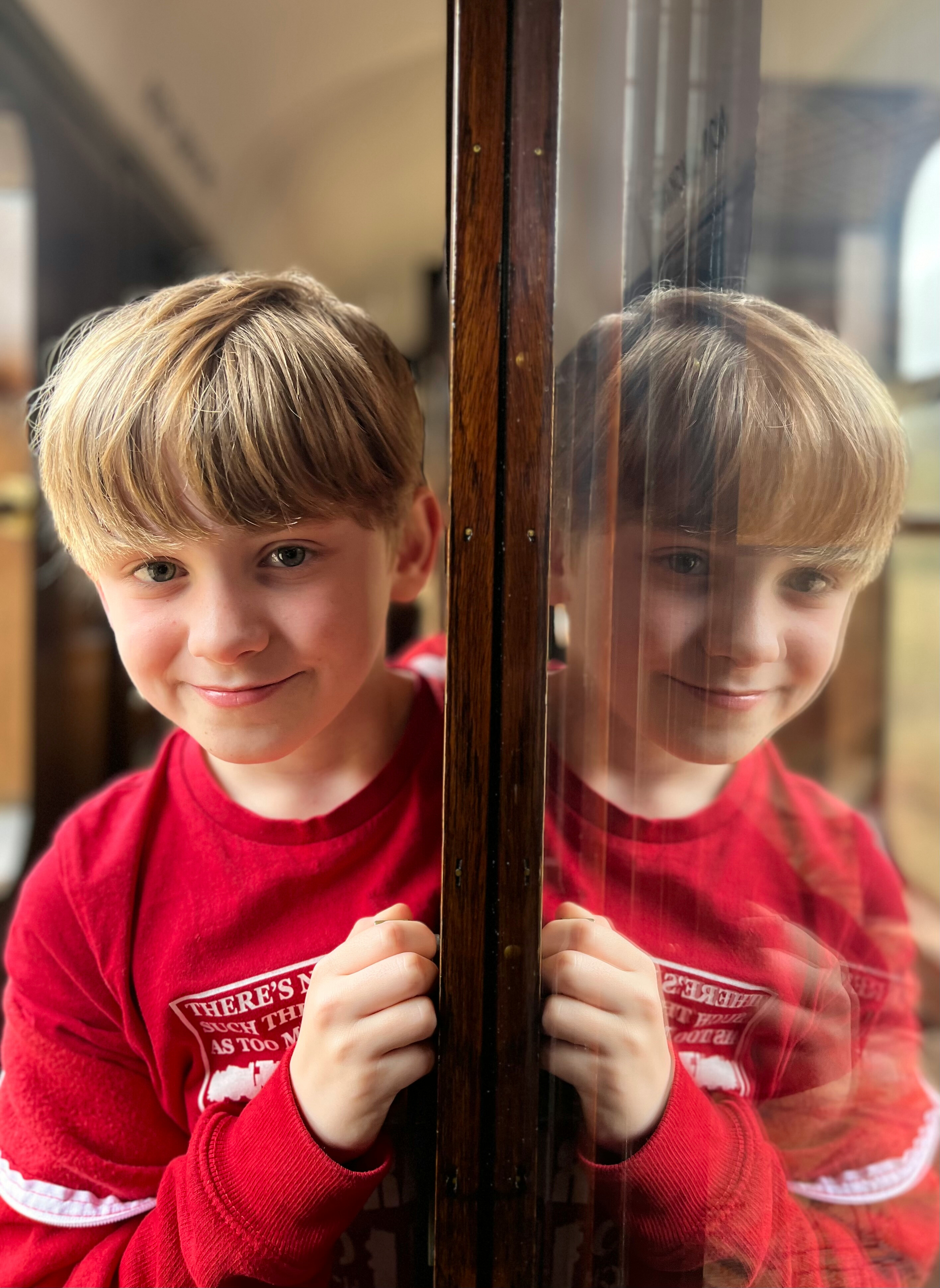 A young rail fan smiles for the camera in NYMR branded red t-shirt, he is reflected in a carriage compartment door.