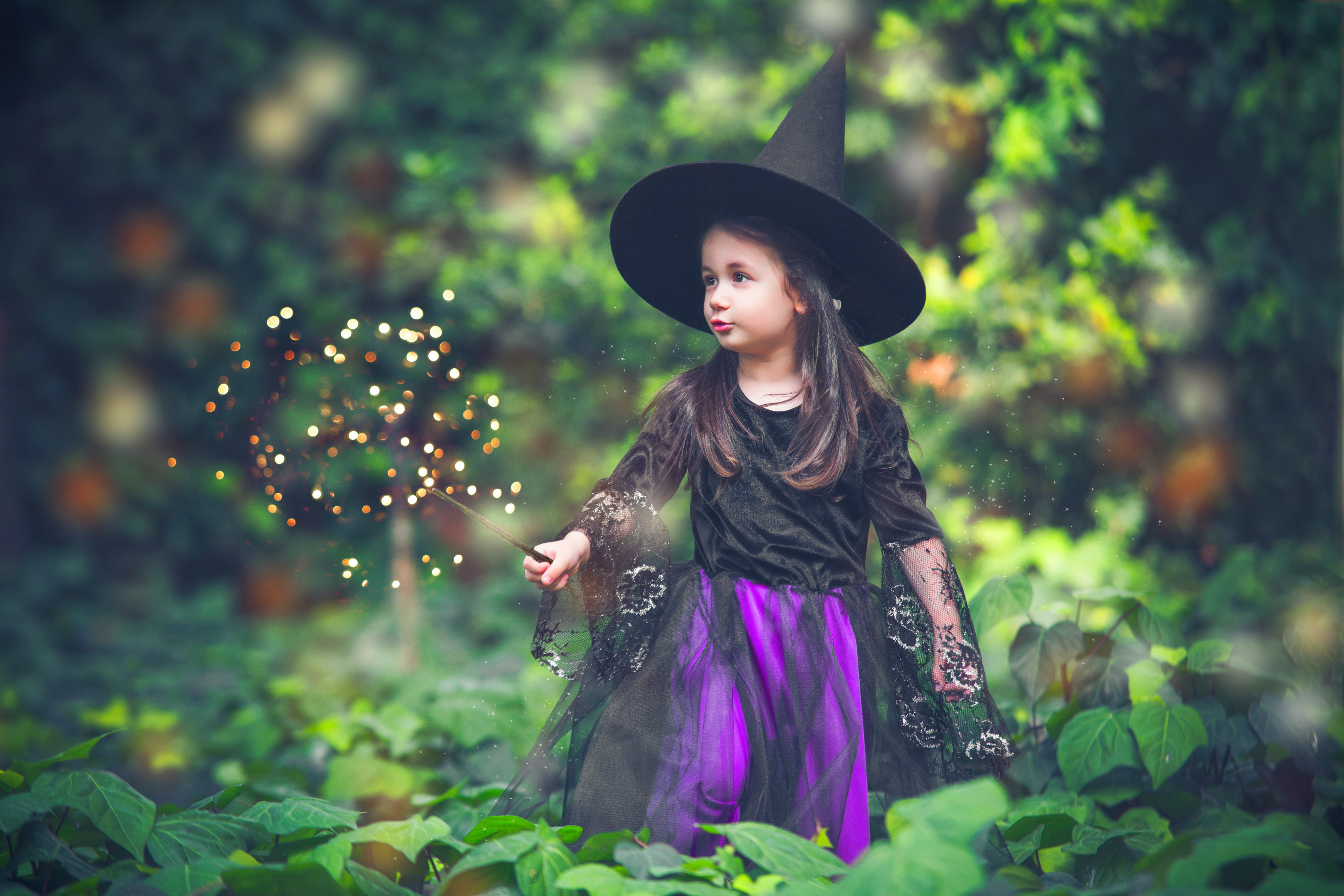 A young girl in a black and purple witches outfit, with wand and witches hat with forest backdrop.