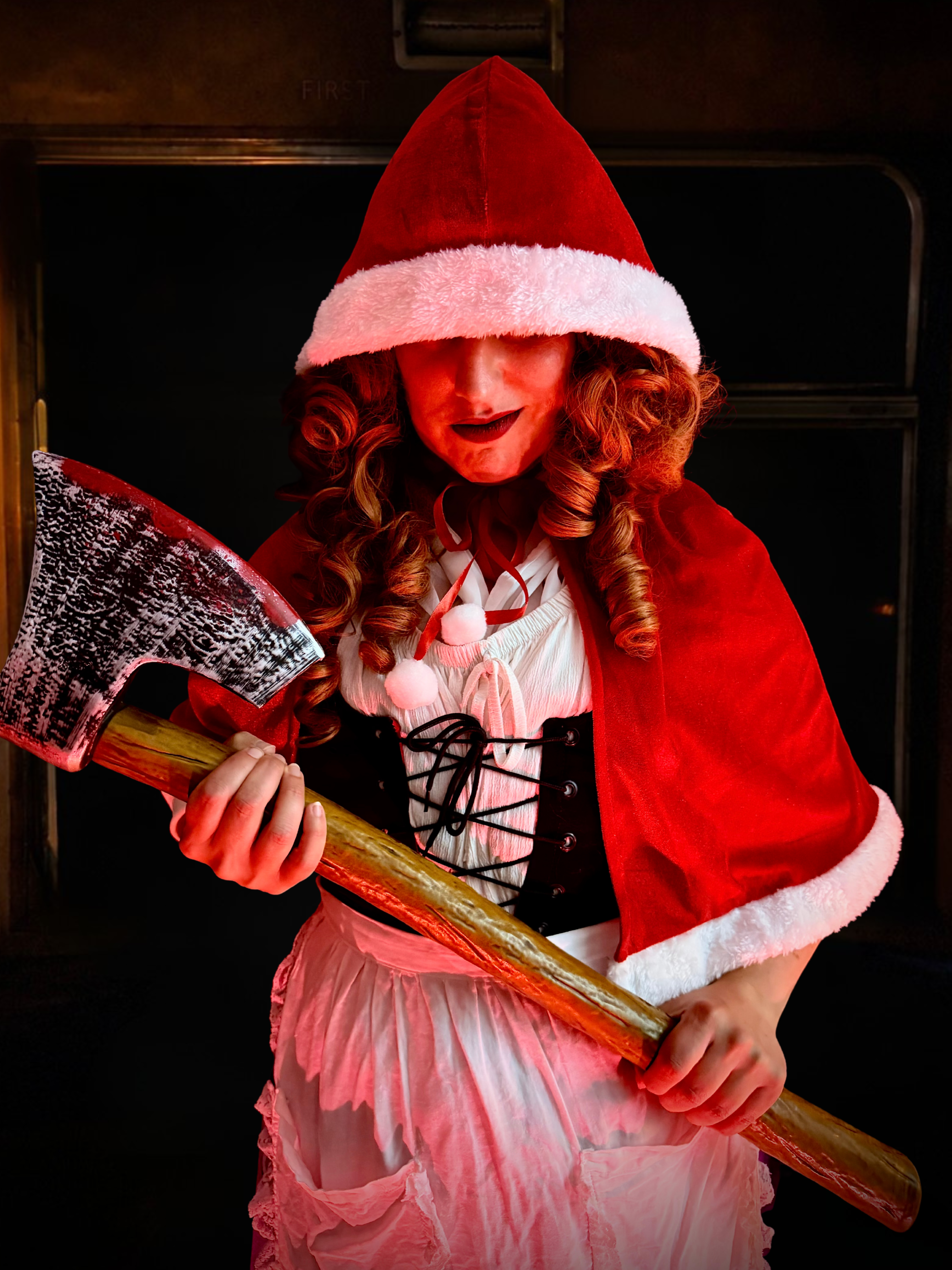 Mad Alice in a red Christmas cape and hood, looking menacing with a bloody axe in hand.