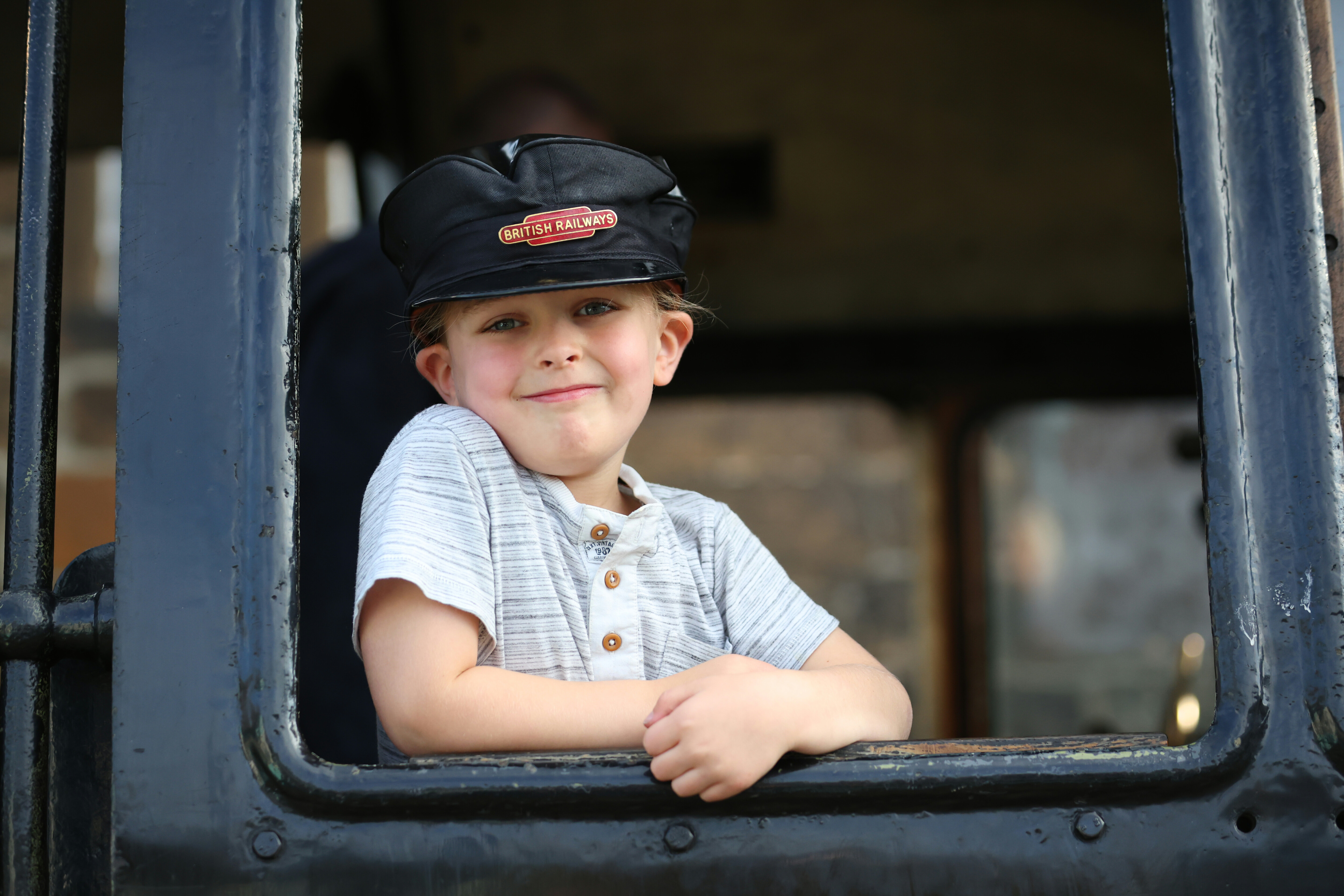 A young girl wears a British Railways drivers hat while posing with a cheeky grin out the footplate window of a locomotive.