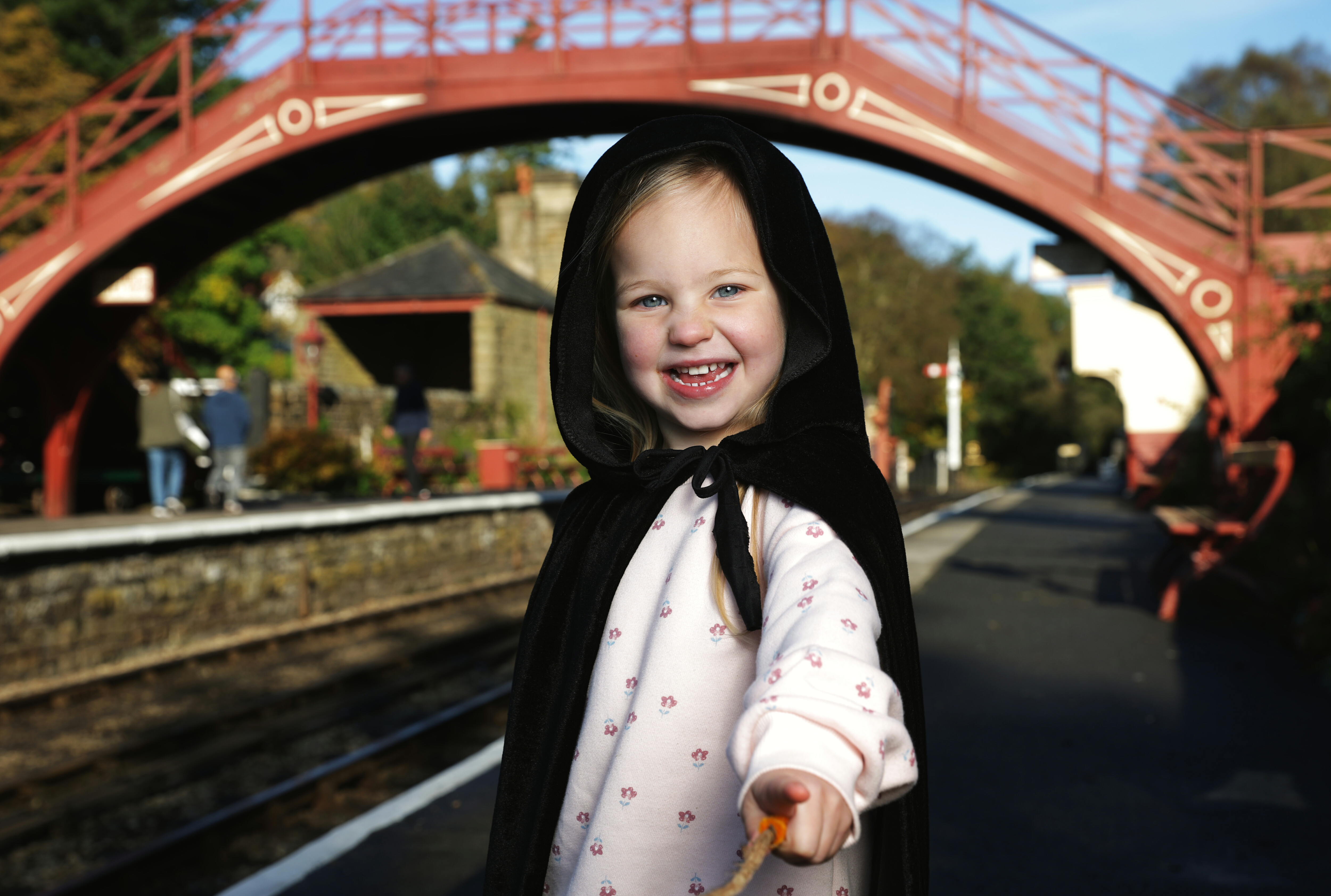 A young girl smiling towards the camera in a black cape, with a wand in hand and Goathland Station as the backdrop.