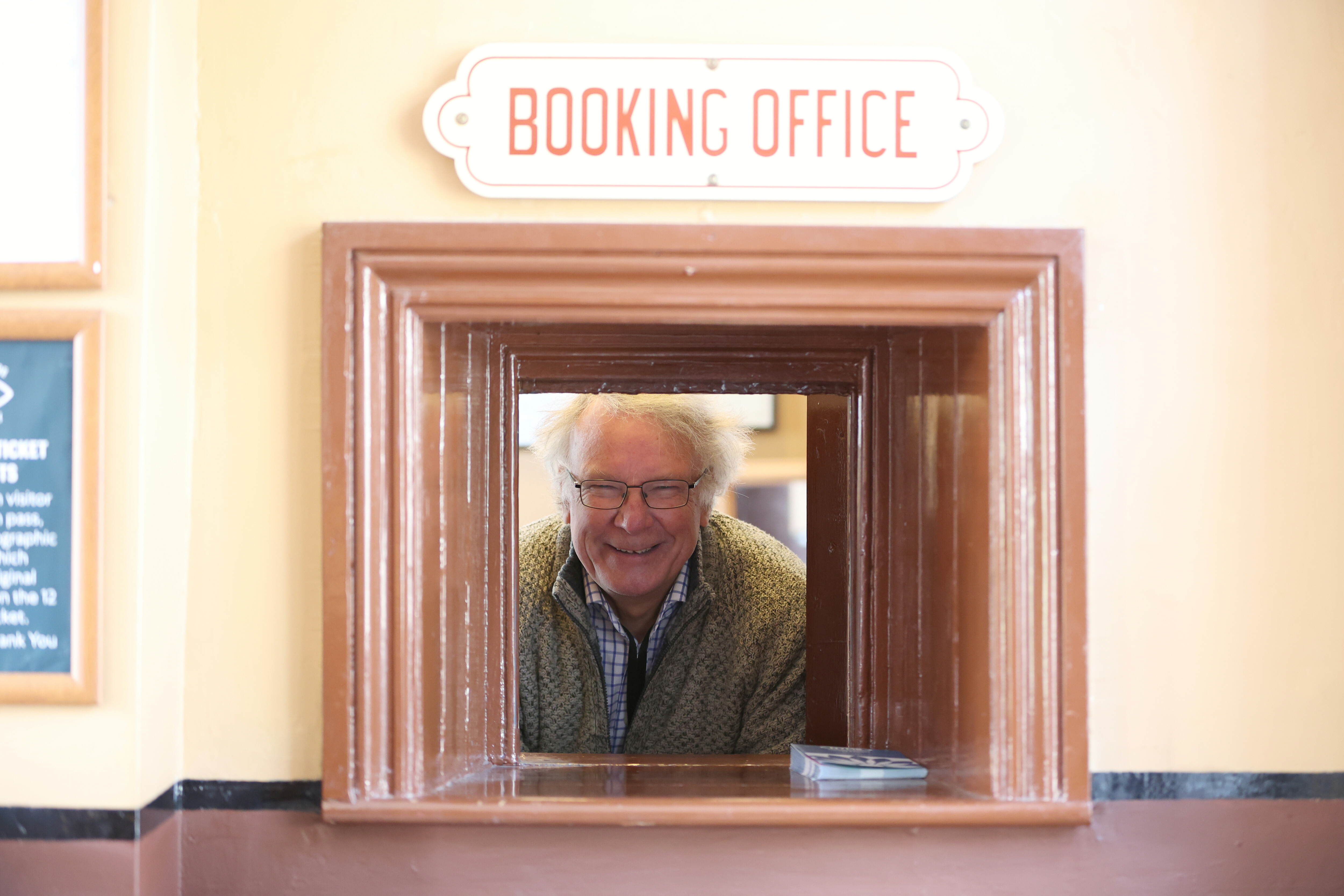A volunteer Booking Office Clerk smiles while looking through the Booking Office window.